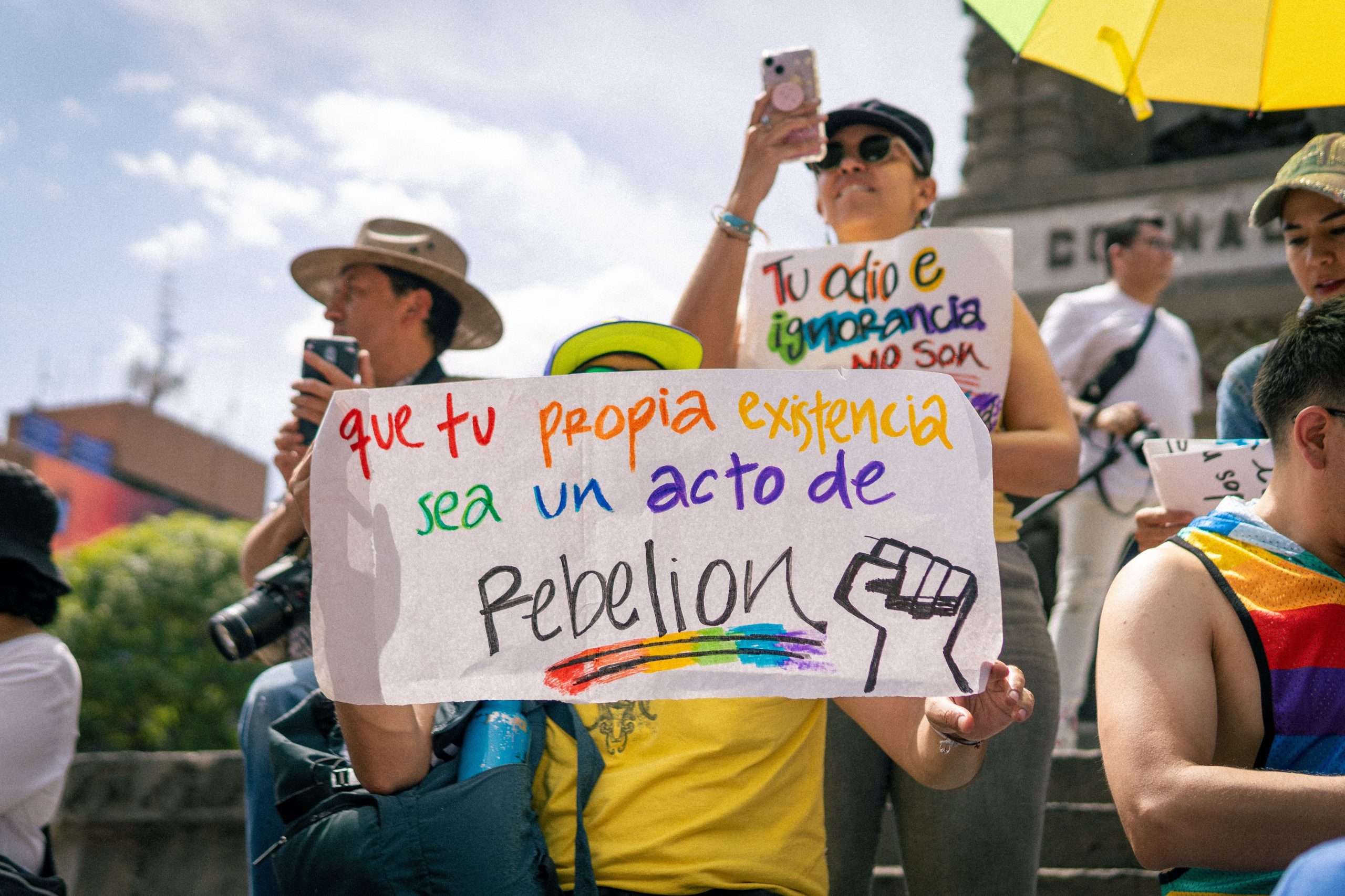 LGBTQ pride event with diverse crowd holding colorful signs and a powerful message of rebellion.
